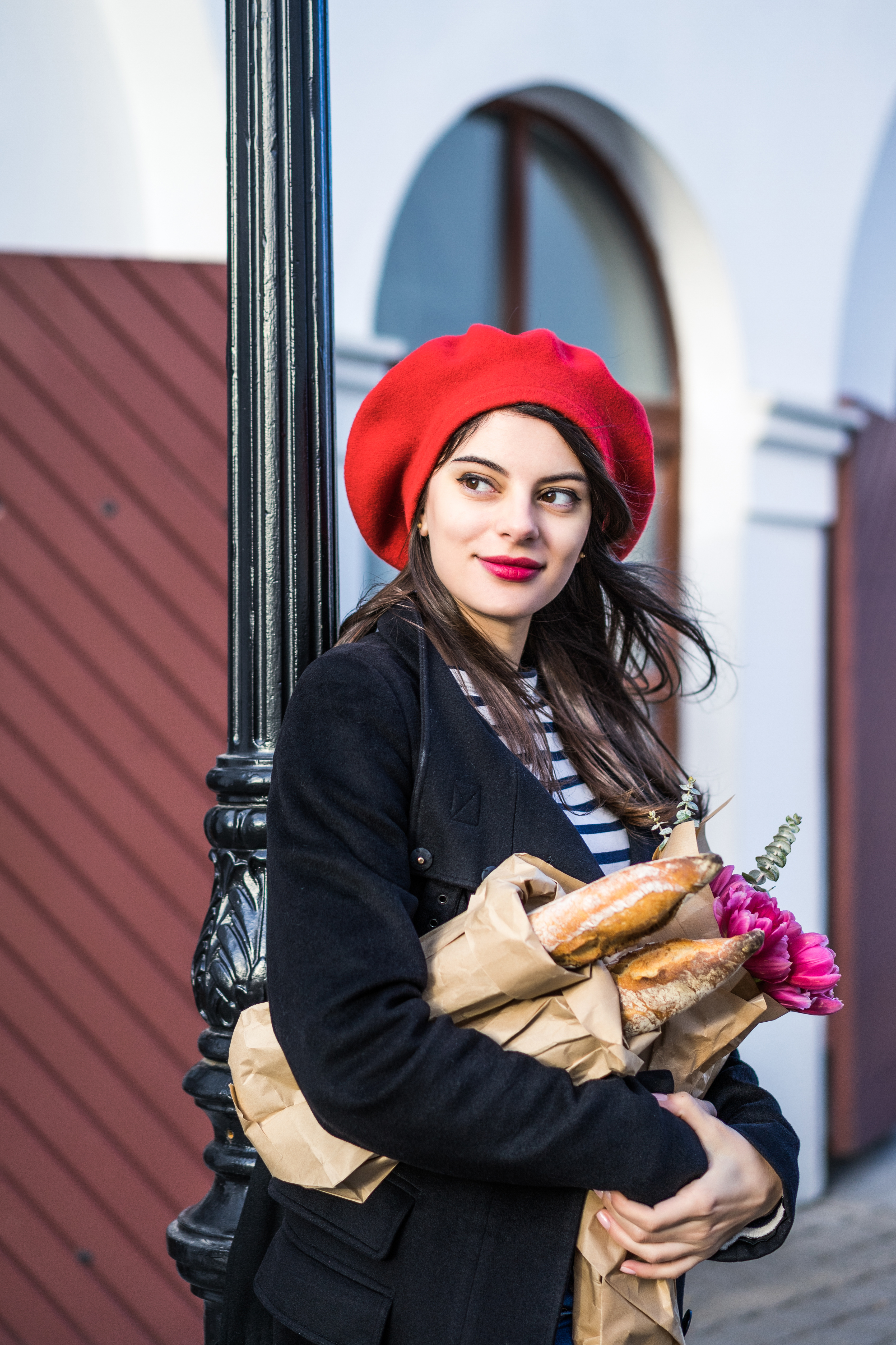 Frenchwoman with baguettes on the street in beret Young beautiful girl Frenchwoman brunette in a red beret and a black coat goes along the street of the European city with a bouquet of flowers and French baguettes. A happy smile and a charming look, the French style of Paris